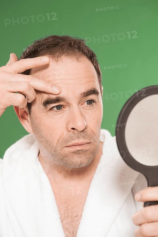 Man wearing a bathrobe observing himself critically in a hand mirror