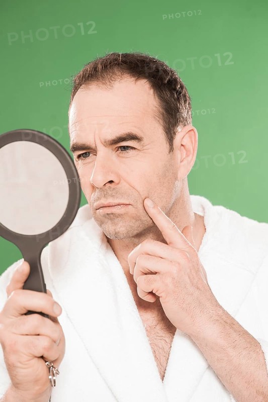 Man wearing a bathrobe observing himself critically in a hand mirror
