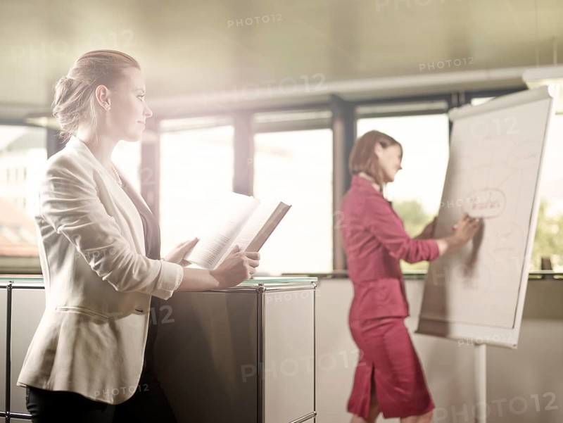 Woman reading a book and looking towards her colleague who is writing on a flip chart