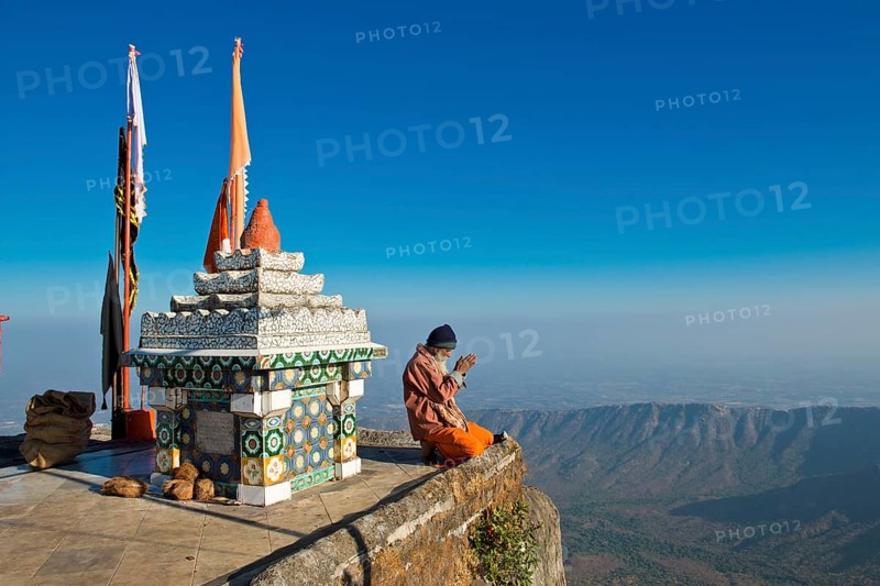 Sadhu or holy man sitting with his hands folded in prayer on the precipice in front of a Hindu temple shrine with prayer flags