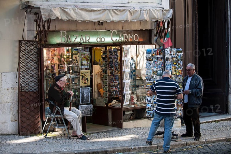Souvenir vendor outside his shop with customers on the Largo do Carmo square