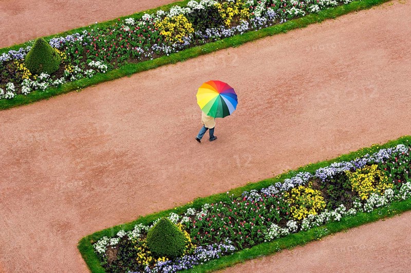 Person with an umbrella in rainbow colours on a red dirt road between flowerbeds in the Palace Gardens