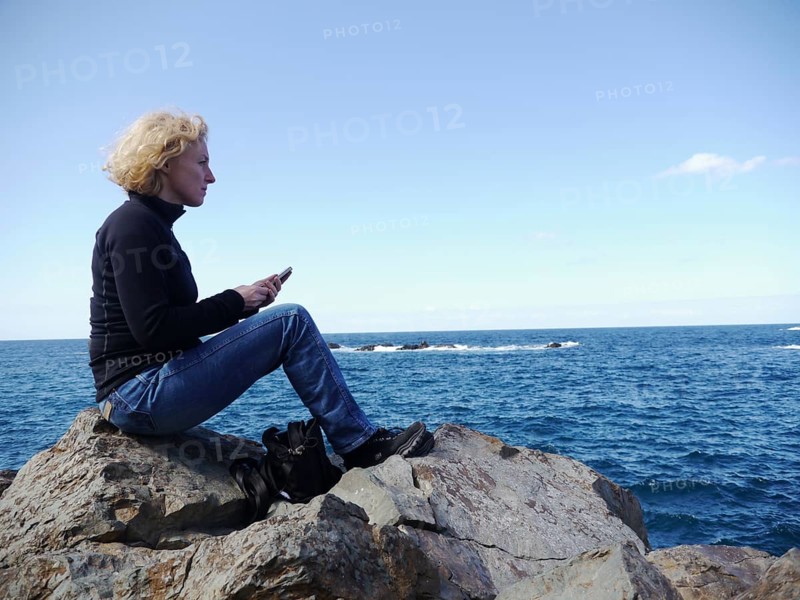 Woman with phone sitting on a rock and looking over the sea