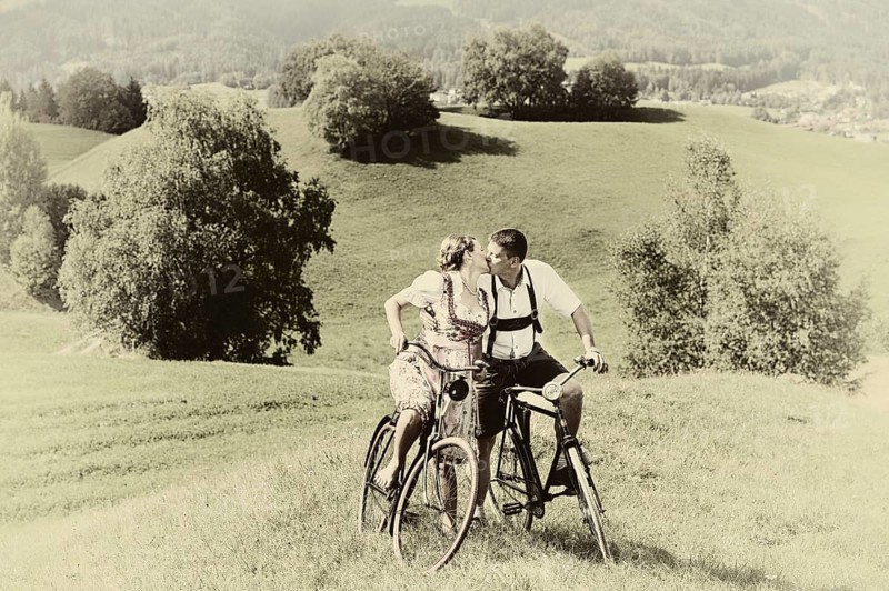 Man wearing leather pants and a woman wearing a dirndl kissing on old bicycles within a natural landscape