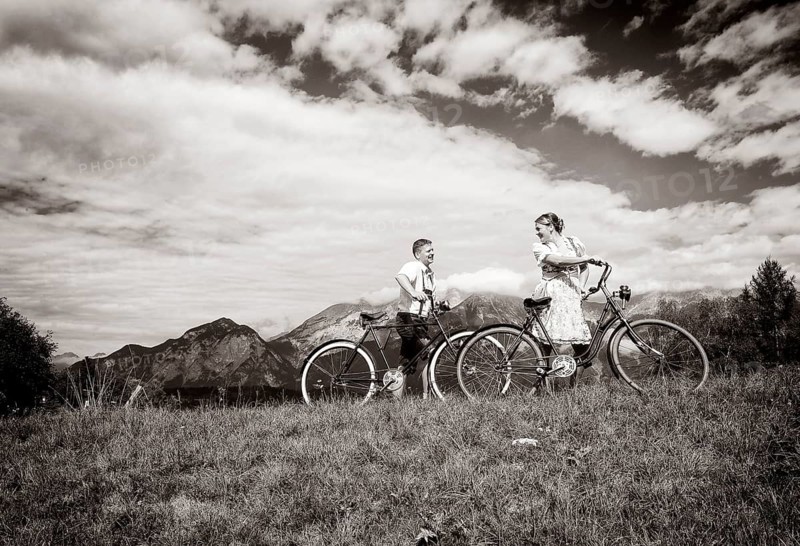 Man wearing leather pants and a woman wearing a dirndl with old bicycles within a natural landscape