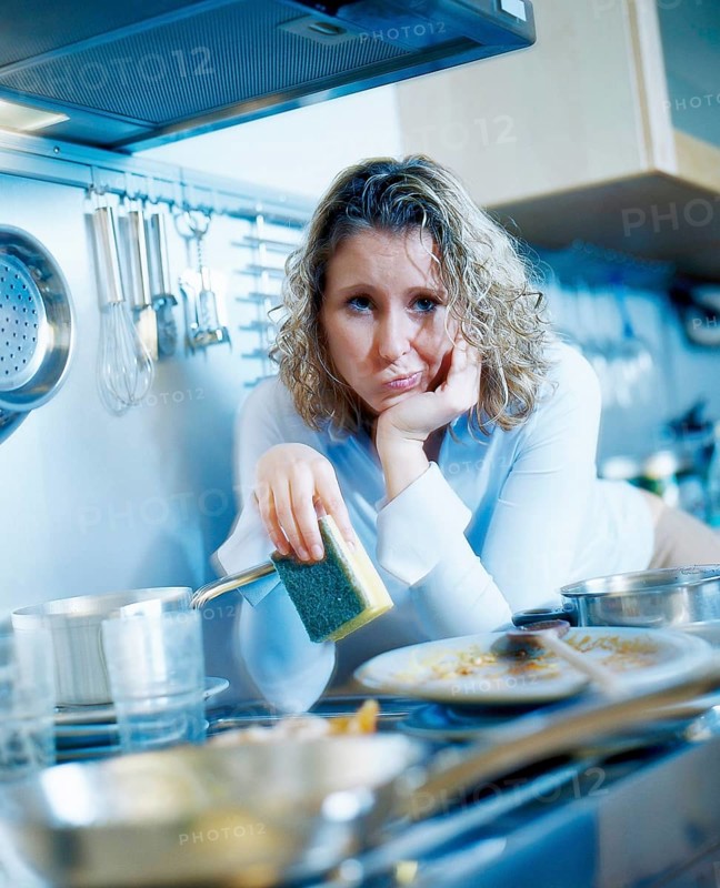 Annoyed woman holding a sponge while standing in a kitchen with dirty dishes