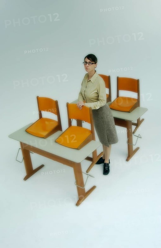 Strict female teacher standing at old school desks with orange chairs