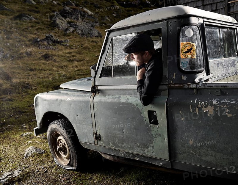 Man with a hat sitting in the driver's seat of an old car noticing a flat front left tyre