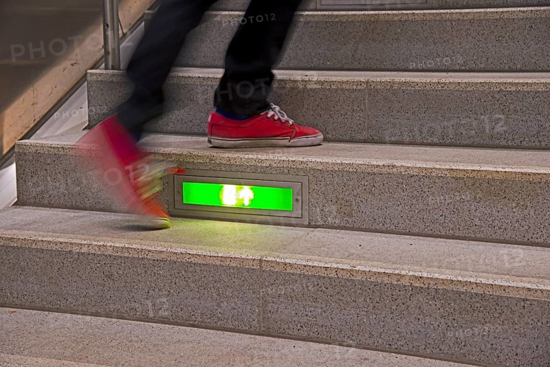 Person rushing up a public staircase with a green illuminated emergency exit sign