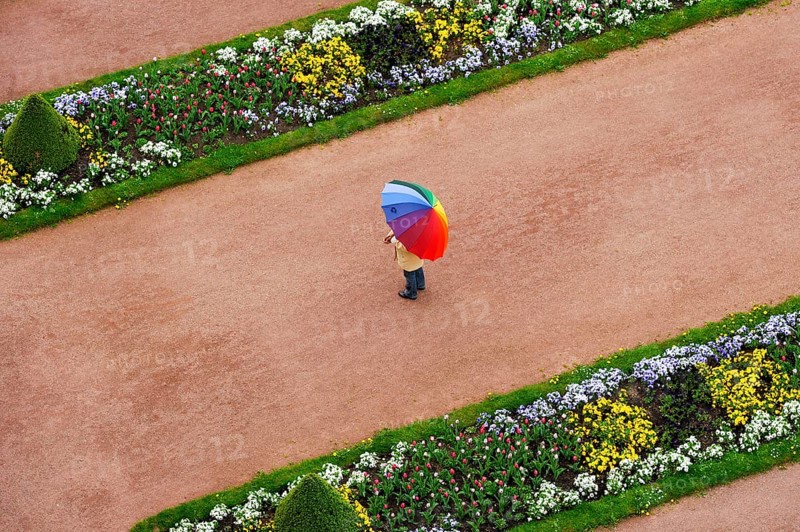Person with umbrella in rainbow colors on a red gravel path between flower beds in the castle gardens