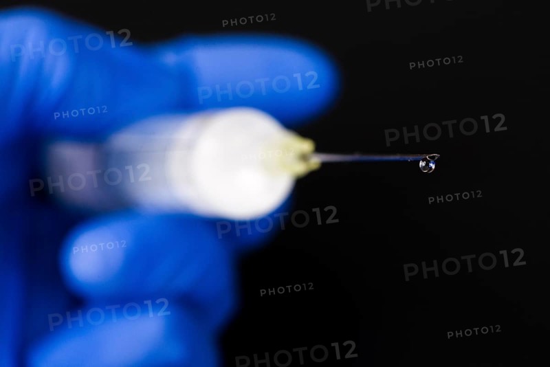A hand with a blue medical glove is holding a syringe with a hypodermic needle and a drop of medicine