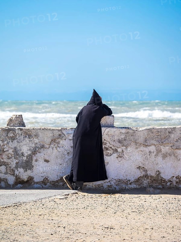 A Berber with black djellaba leaning against the harbor wall