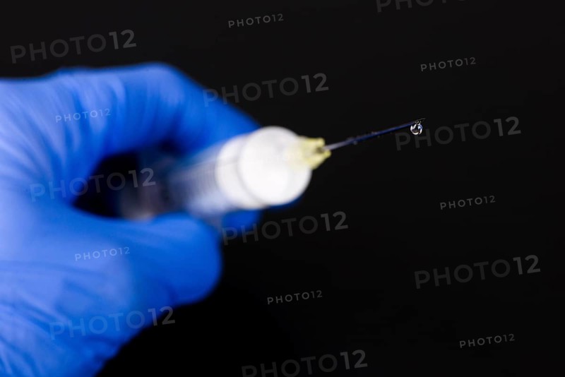 A hand with a blue medical glove is holding a syringe with a hypodermic needle and a drop of medicine