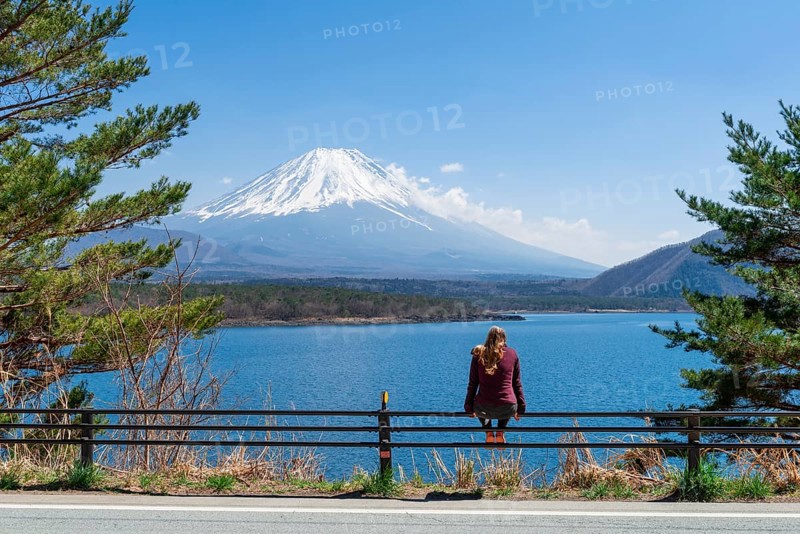 Young woman sitting on a railing next to a road and looking across the lake to the volcano Mt Fuji