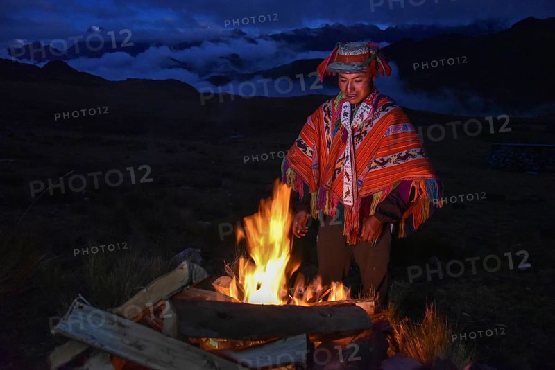Indio man with hat and colorful traditional poncho standing at a campfire in the Andes