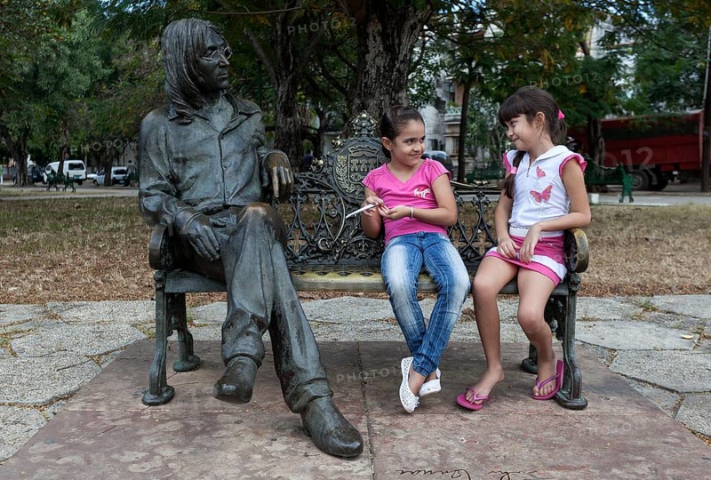 Two girls sitting on a bench next to a bronze sculpture of John Lennon