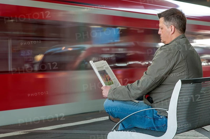 Man sitting on a bench at a station reading a newspaper