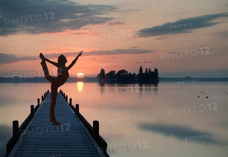 Woman with backlighting doing a yoga pose on a wooden pier at a lake in front of the setting sun