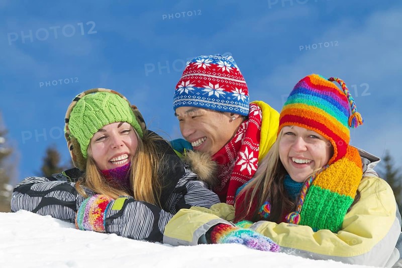 Young Asian man and two young European women smiling while lying on a mound of snow and wearing colourful winter clothing