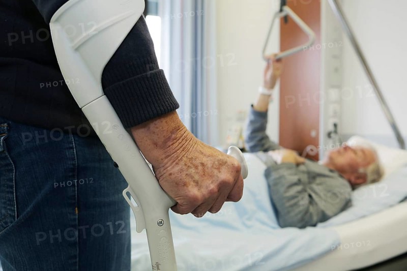 Elderly woman standing on a crutch in front of an elderly man lying in a hospital bed