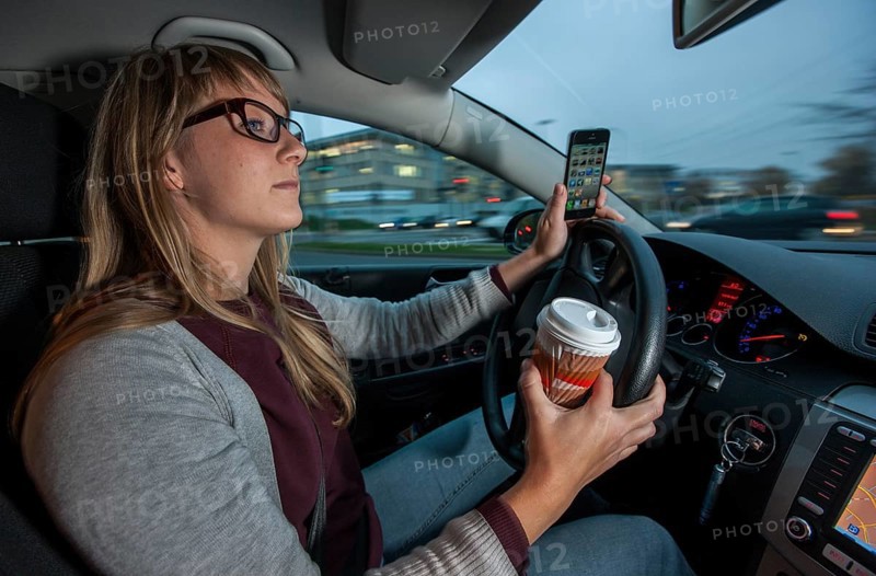 Young woman driving a car in the city centre while holding her cell phone and a cup of coffee at the same time