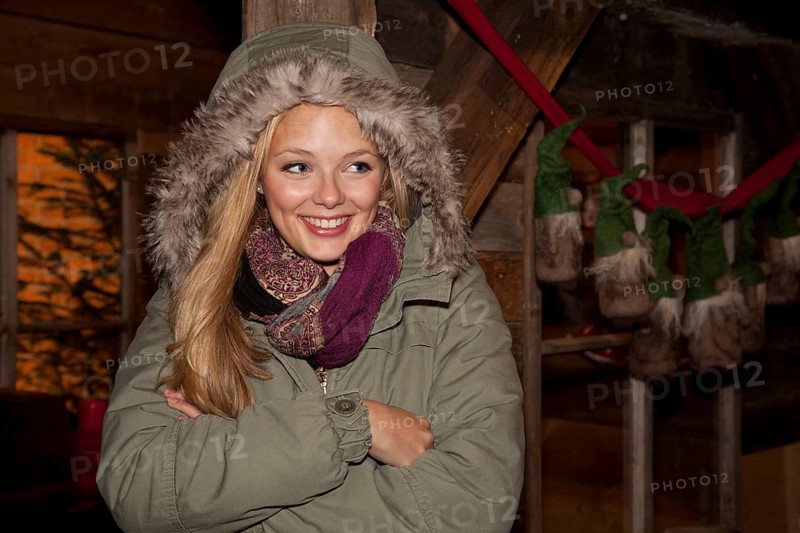 Smiling young woman wearing a green parka standing with her arms crossed in front of a hiking hut decorated for Christmas