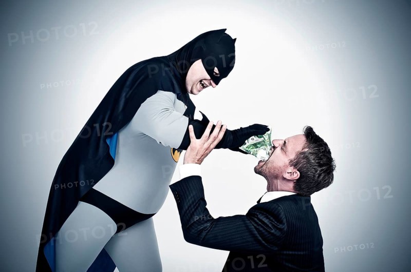 Chubby superhero wearing a Batman costume stuffing money into the mouth of a man wearing a suit