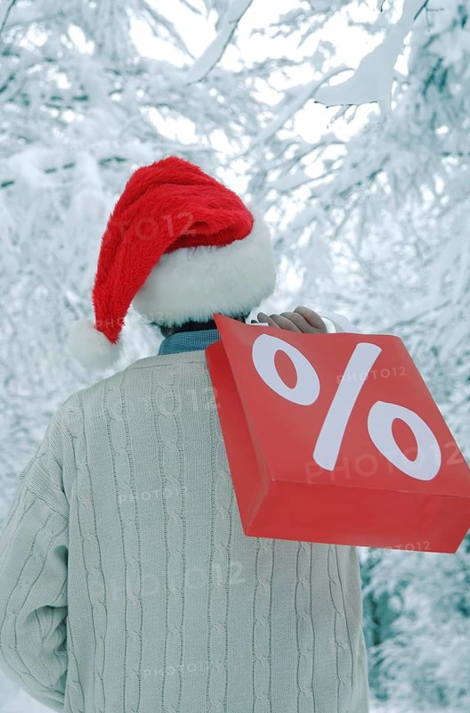 Man wearing a Santa hat in a snow-covered forest holding a shopping bag with a percent sign