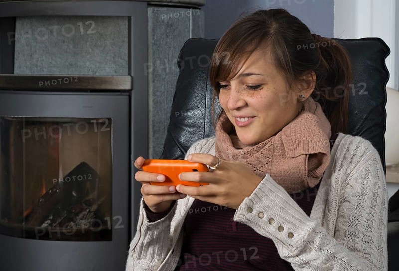 A young woman of Indonesian origin is sitting in front of a wood burner working on a smartphone