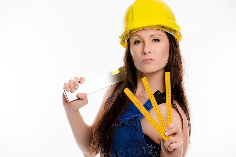 Woman wearing blue overalls and a hardhat holding a folding carpenter's ruler and a spirit level