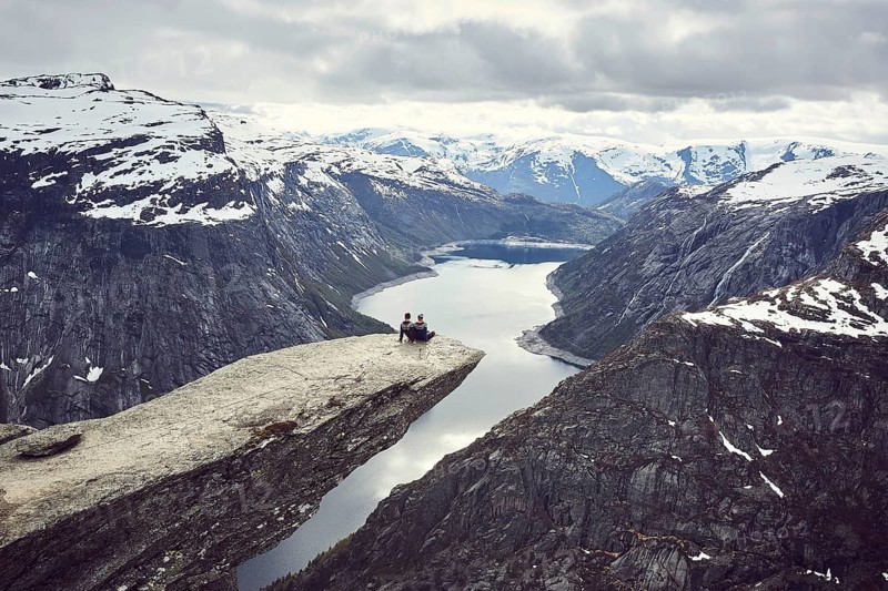 Paar sits on a rock platform on Trolltunga and looks over fjord landscape