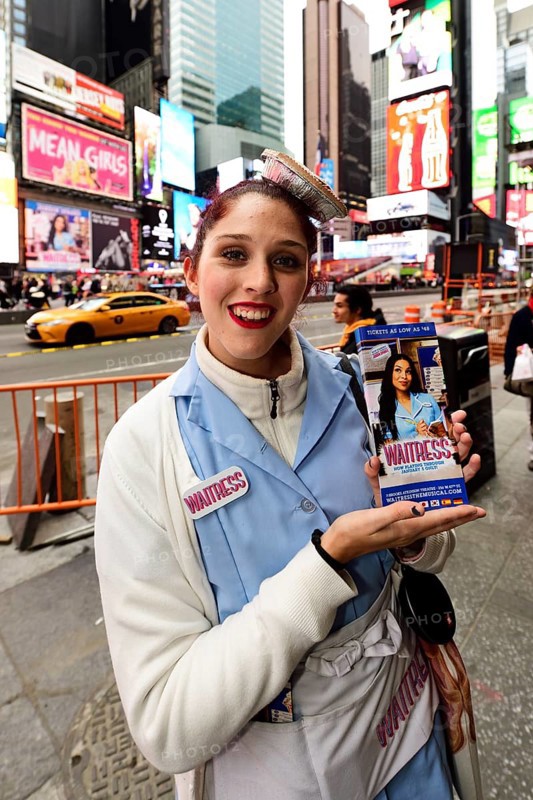 Young woman in waitress costume and cupcakes on her head promoting a musical