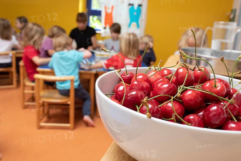 A bowl of cherries stands in the dining room in the kindergarten and children sit at the table to eat