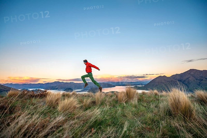 Hiker jumps into the air, panoramic view of Wanaka Lake and mountains at sunset