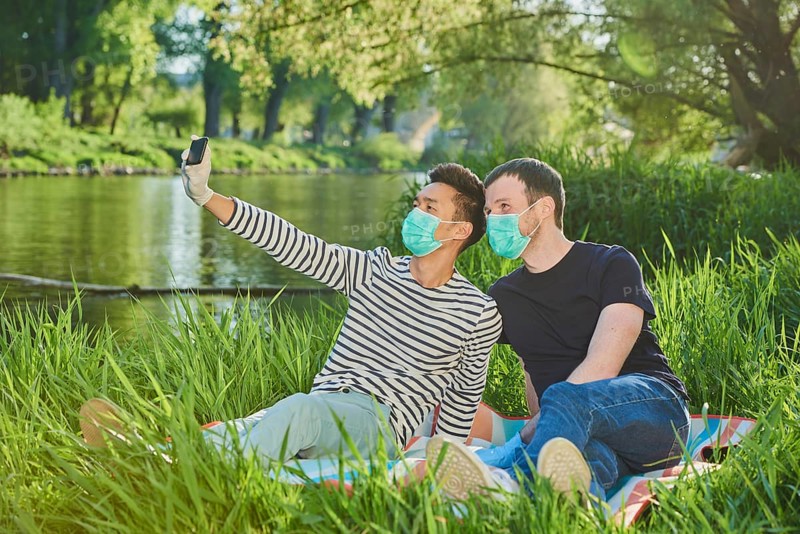 Two homosexual men with face masks sitting in a meadow by the river