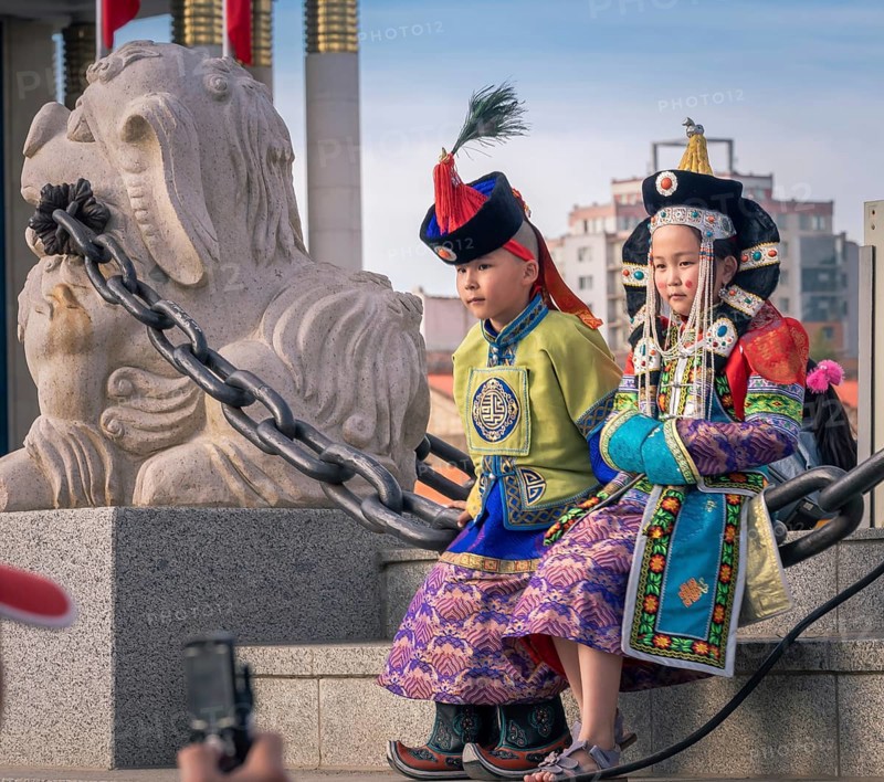 Mongolian boy in the traditional traditional traditional traditional traditional traditional traditional traditional traditional traditional costume of a man and a girl in the traditional costume of a woman sitting on a chain next to the lion statue in the central square