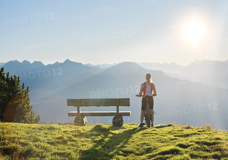 Mountain biker stands with MTB next to a wooden bench and looks into the wide light-flooded mountain landscape