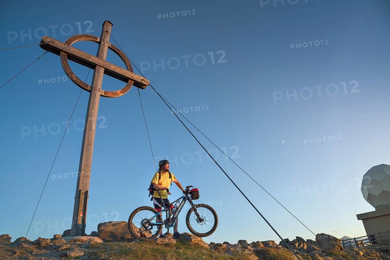 Mountain biker stands with e-MTB next to the summit cross of the Patscherkofel in the warm evening light