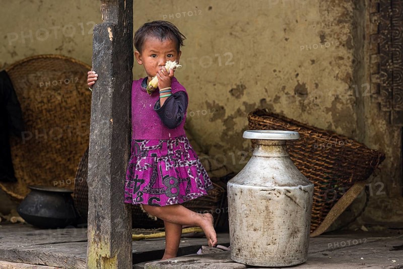 A little girl stands in front of the house next to an aluminium water container and chews sugar cane
