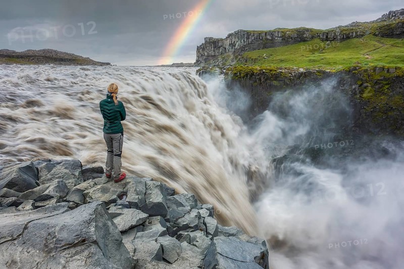 Blond woman with green down jacket standing at Dettifoss with rainbow