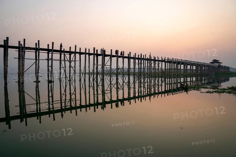 Buddhist monks walk across the U-leg bridge in red robes as the sun rises