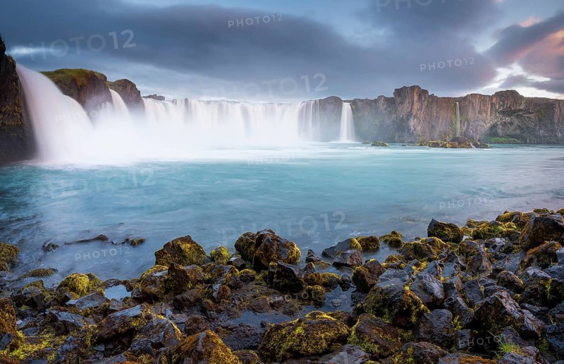Long time exposure of a turquoise waterfall in volcanic landscape with dramatic clouds and green moss on rocks