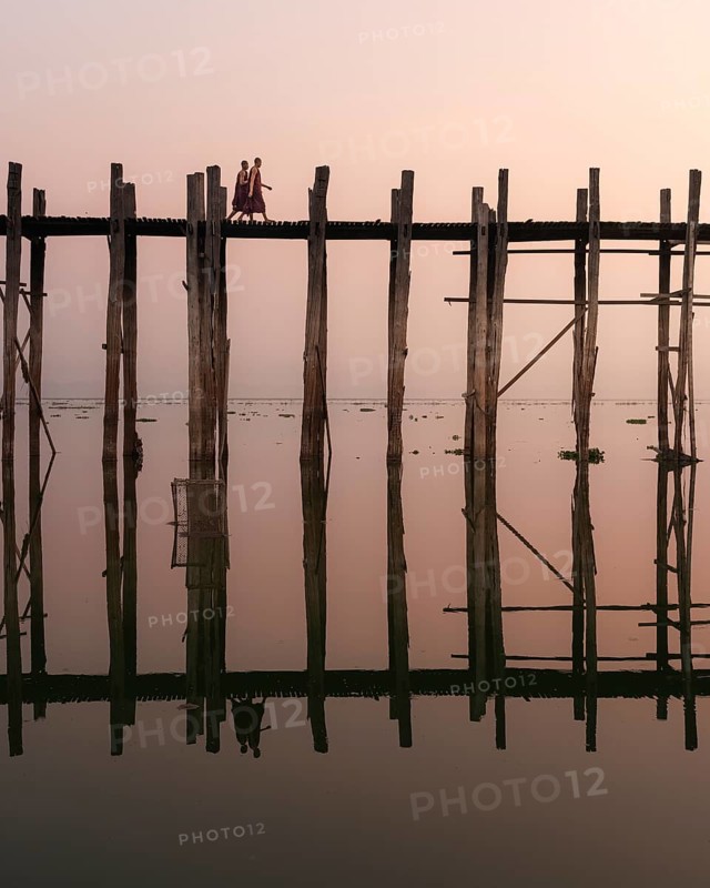 Buddhist monks walk across the U-leg bridge in red robes as the sun rises