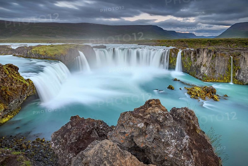 Long time exposure of a turquoise waterfall in volcanic landscape with dramatic clouds