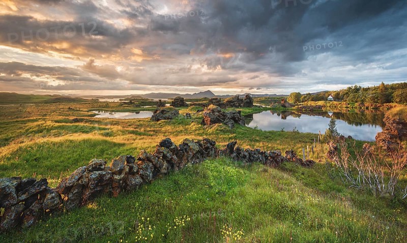 Stone wall and lava formations of volcanic rock rise from green meadow with dramatic sky in the evening light at Kalfastroend