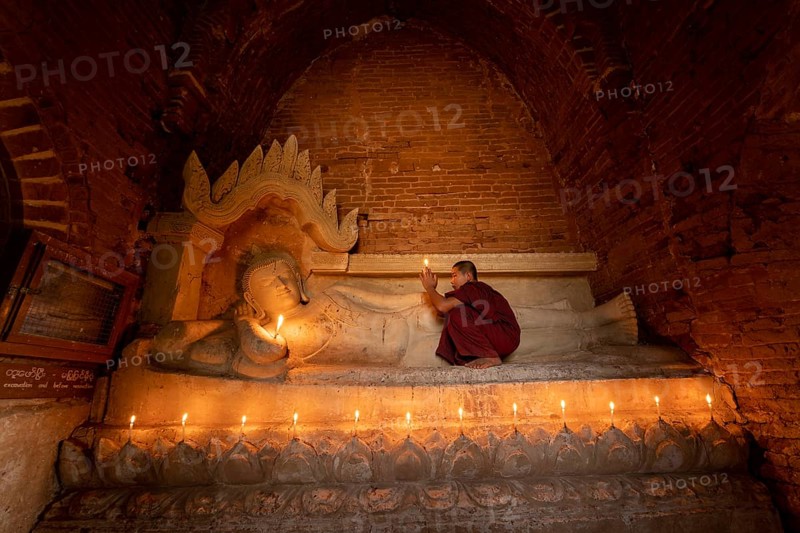 Buddhist young monk in red robe with red umbrella praying with candle in hand in a temple