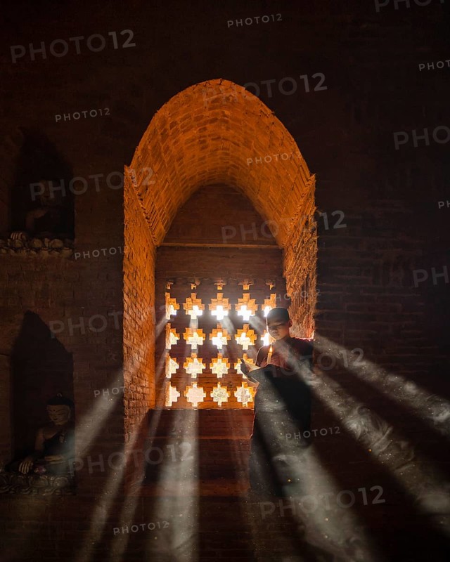 Buddhist young monk in red robe reading standing in front of rays of light in a temple