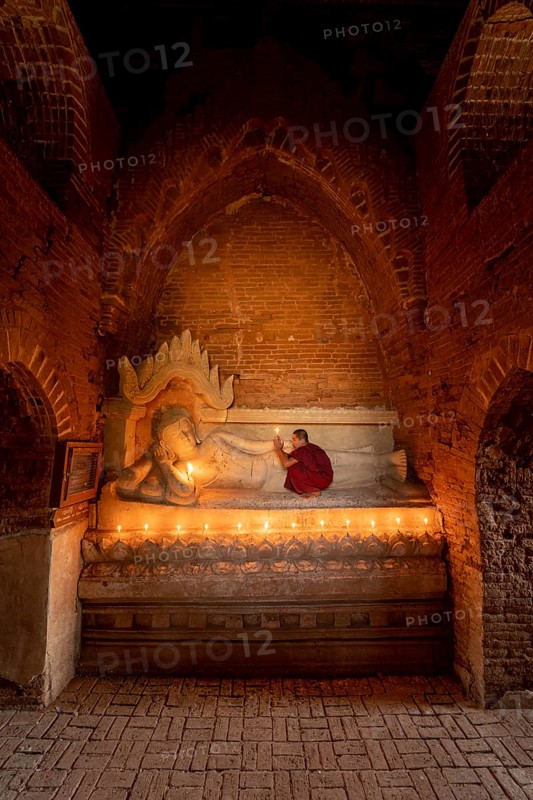 Buddhist young monk in red robe with red umbrella praying with candle in hand in a temple