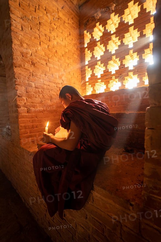 Buddhist young monk in red robe reading while sitting with a candle in front of rays of light in a temple