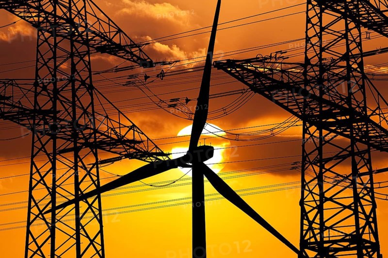 Two overhead line pylons and a wind turbine in front of the sun